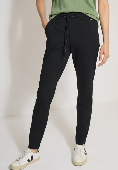 Spodnie Mid Waist z wąskimi nogawkami o Casual Fit Black Kobieta w czarnych dresowych spodniach z sznurkiem, w białych trampkach na neutralnym tle.