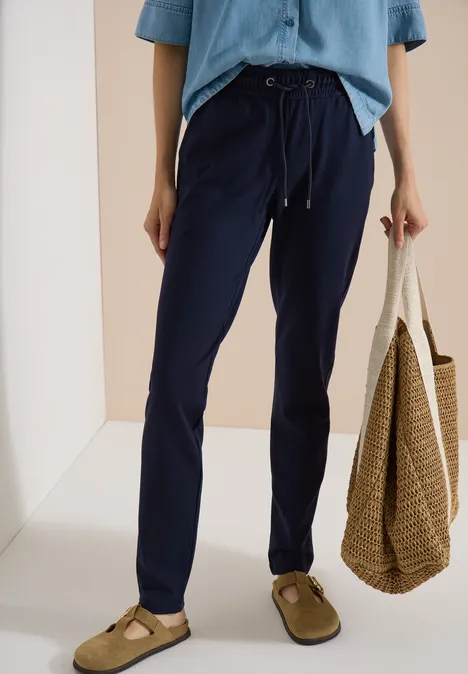 Mid Waist Slim Leg Hose in casual pasvorm urban dark blue Vrouw draagt donkerblauwe vrijetijdshose met tunnelkoord, lichtblauwe blouse, strooien tas en instappers.