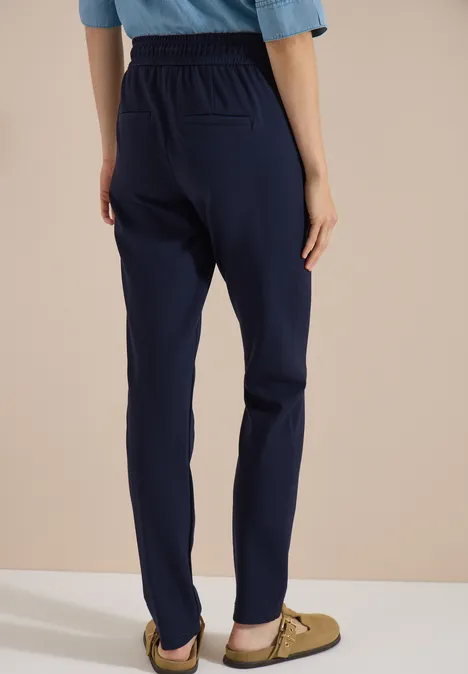 Mid Waist Slim Leg Hose in casual pasvorm urban dark blue Persoon van achteren in rechte donkerblauwe broek met elastische taille en beige loafers tegen lichte achtergrond.