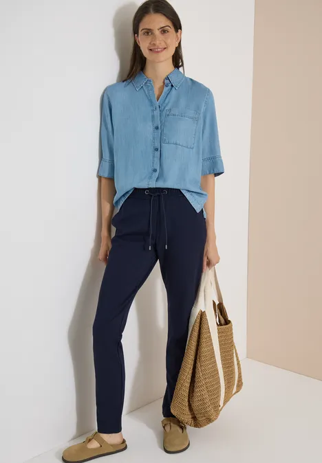 Mid Waist Slim Leg Hose in casual pasvorm urban dark blue Vrouw in blauw overhemd en donkerblauwe broek met grote strotas voor een lichte achtergrond.