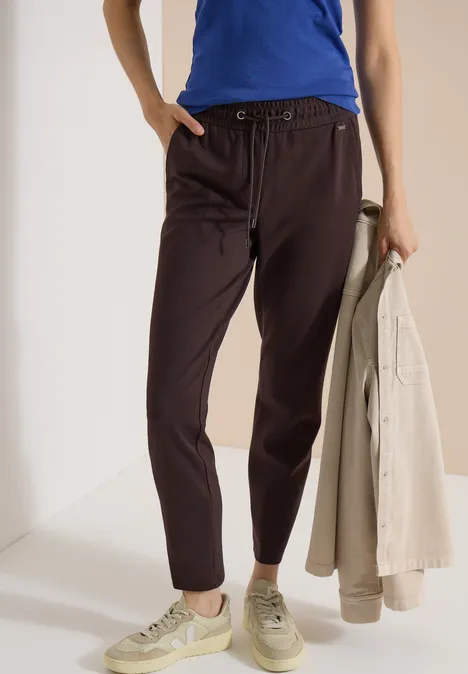 Mid Waist Slim Leg Hose in casual pasvorm truffle brown Vrouw draagt blauw shirt, donkerbruine broek en lichte sneakers, houdt een beige jasje in haar hand.