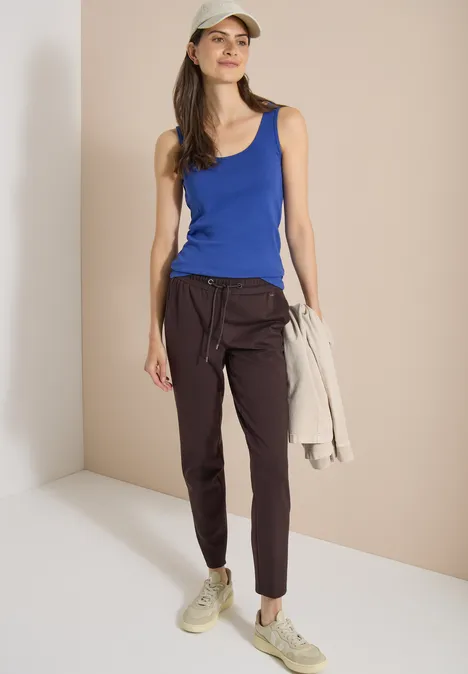 Mid Waist Slim Leg Hose in casual pasvorm truffle brown Een vrouw in een blauw tanktop, donkere broek en sneakers houdt een licht jasje vast.