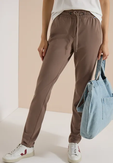 Vrouw in bruine joggerbroek met trekkoord, witte sneakers en grote blauwe denimtas.