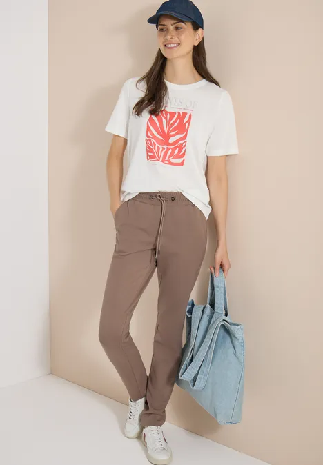 Vrouw met wit t-shirt met rode print, bruine broek, witte sneakers en grote blauwe tas.