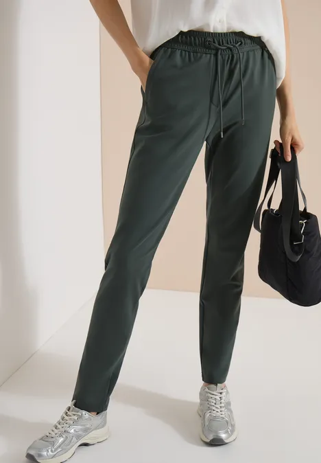 Mid Waist Slim Leg Hose im Casual Fit easy khaki Person trägt dunkelgrüne Jogginghose, weiße Bluse, Sneaker und hält eine schwarze Tasche.