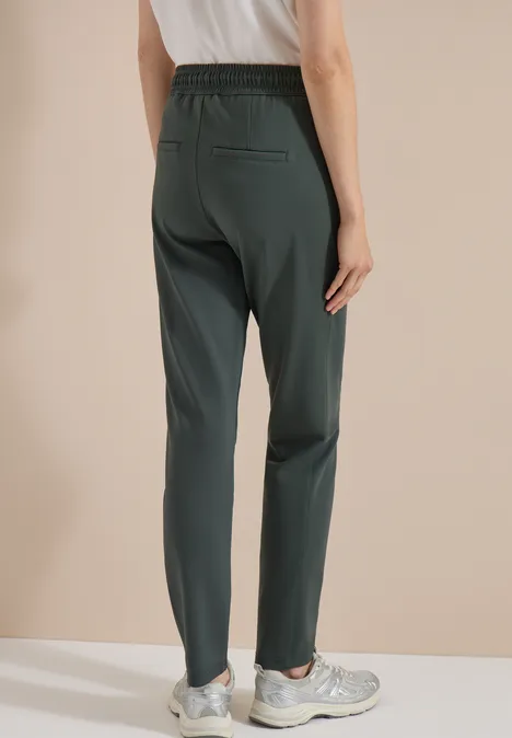 Mid Waist Slim Leg Hose im Casual Fit easy khaki Model trägt dunkle, geradlinige Hose mit elastischem Bund und Sneaker, Ansicht von hinten.