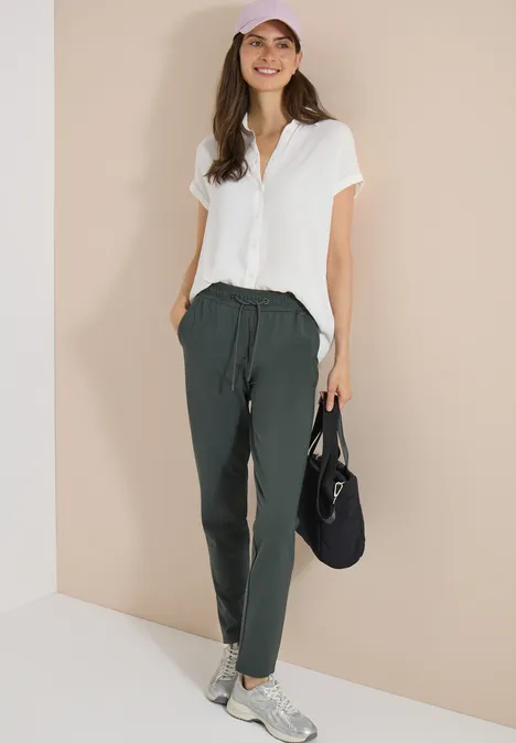 Mid Waist Slim Leg Hose im Casual Fit easy khaki Frau in weißer Bluse und dunkler Jogginghose mit Tunnelzug, weißen Sneakern und schwarzer Tasche vor beigem Hintergrund.