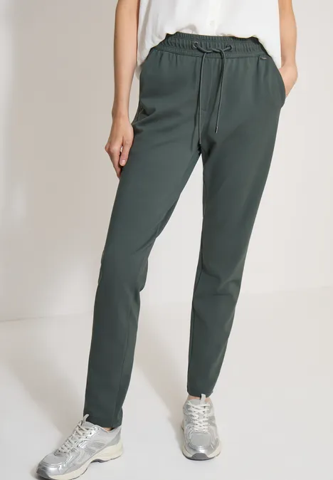 Mid Waist Slim Leg Hose im Casual Fit easy khaki Modell trägt dunkelgrüne Hose mit Tunnelzug und silbernen Sneakern.