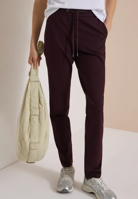 Person trägt eine weiße Bluse, bordeauxrote Hose, graue Sneaker und hält eine hellbeige Umhängetasche.