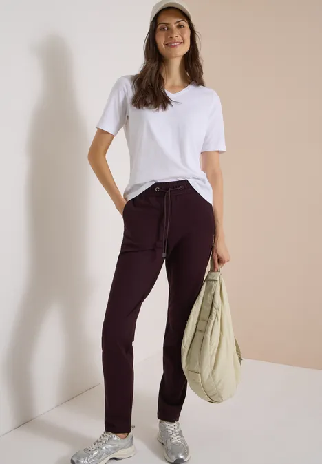 Frau trägt weißes T-Shirt, weinrote Hose, Baseball-Cap und hält eine beige Tasche vor hellem Hintergrund.