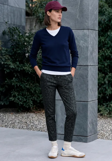 Une femme appuyée sur une colonne, en pantalon gris à motif, pull bleu marine, T-shirt blanc, baskets et casquette bordeaux.