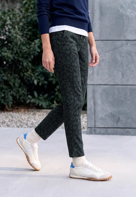 Personne portant un pantalon noir à motif et des baskets blanches sur un sol clair devant un mur et des buissons.