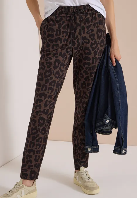 Mid Waist Slim Leg Hose im Casual Fit dark toffee brown Frau in locker sitzender, braun-schwarzer Hose mit Leopardenmuster und Sneakern, hält Jeansjacke in der Hand.