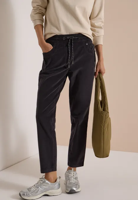 Mid Waist Slim Leg Hose im Casual Fit carbon grey Person trägt schwarze Cordhose, hellen Pullover und silberne Sneaker, hält eine olivgrüne Tasche.