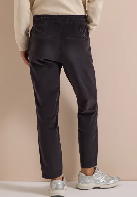 Mid Waist Slim Leg Hose im Casual Fit carbon grey Rückenansicht einer Person mit dunkler Hose und silbernen Sneakern vor neutralem Hintergrund.