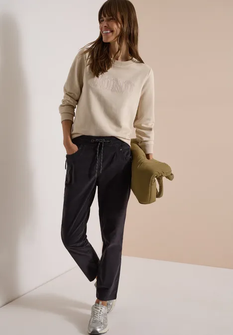 Mid Waist Slim Leg Hose im Casual Fit carbon grey Frau mit beige Sweatshirt, schwarzen Jogginghosen, silbernen Sneakern und olivgrünem Mantel vor hellem Hintergrund.