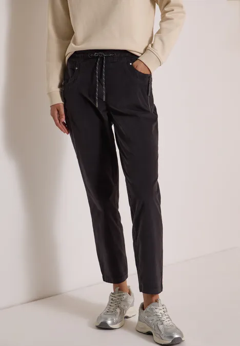 Mid Waist Slim Leg Hose im Casual Fit carbon grey Schwarze Hose mit Tunnelzug, schmaler Schnitt, kombiniert mit hellbeigem Pullover und grauen Sneakern.