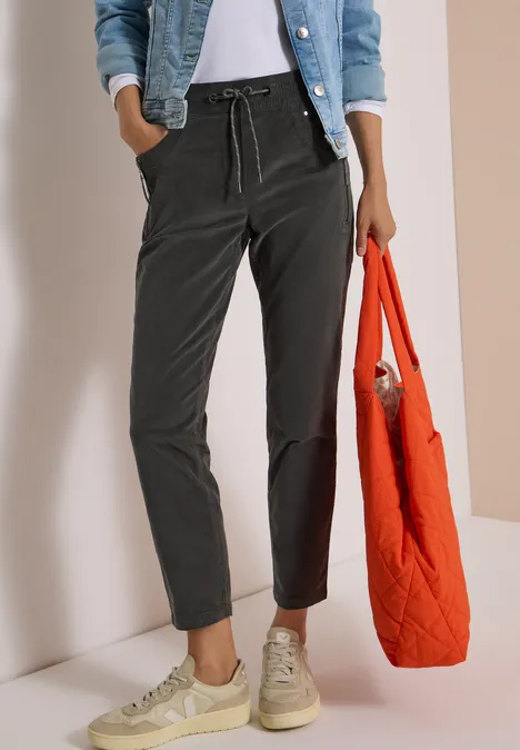 Mid Waist Slim Leg Hose im Casual Fit easy khaki Frau in schwarzer Hose, Jeansjacke, weißen Sneakern und Top, mit orangefarbenem Stepp-Shopper.