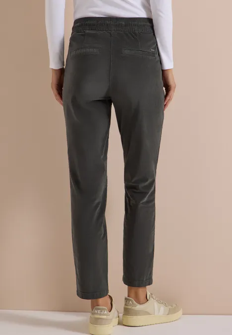 Mid Waist Slim Leg Hose im Casual Fit easy khaki Rückansicht einer Person in dunkelgrauen, lässig sitzenden Hosen und hellen Sneakern vor beigem Hintergrund.