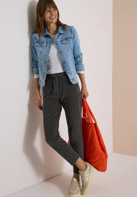 Mid Waist Slim Leg Hose im Casual Fit easy khaki Frau in Jeansjacke, grauem Shirt und schwarzen Jogginghosen lächelt, hält rote Tasche.