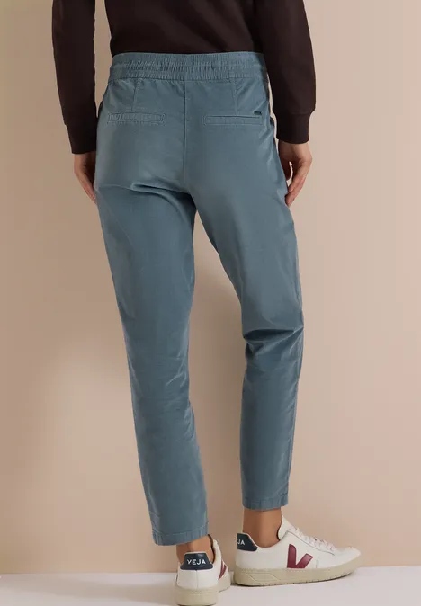 Mid Waist Slim Leg Hose im Casual Fit dove blue Taillierte, türkise Hose mit elastischem Bund und Taschen, kombiniert mit weißen Sneakern vor beigem Hintergrund.