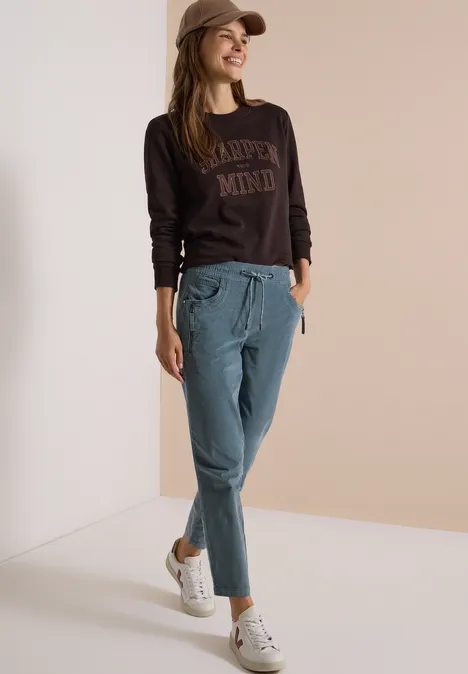 Mid Waist Slim Leg Hose im Casual Fit dove blue Frau mit Cap trägt braunes Sweatshirt, blaue Chinohose und weiße Sneaker, lässig vor hellem Hintergrund.