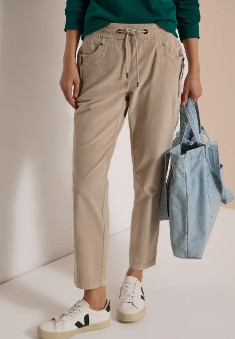 Mid Waist Slim Leg Hose im Casual Fit silvery beige Beige Bundfaltenhose, weiße Sneaker und blaue Stofftasche an der Hand.