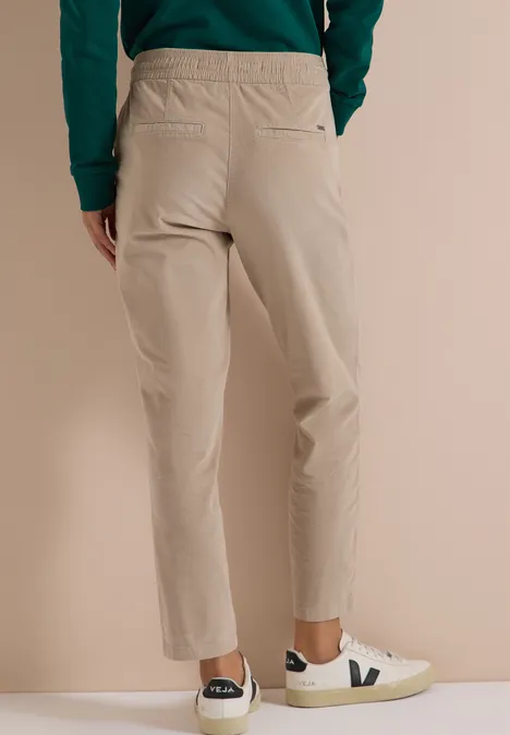 Mid Waist Slim Leg Hose im Casual Fit silvery beige Rückansicht einer Person in beiger Hose und weißen Sneakers mit V-Logo vor neutralem Hintergrund.