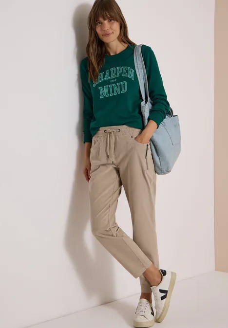 Mid Waist Slim Leg Hose im Casual Fit silvery beige Frau in grünem Sweatshirt und beigen Hosen mit hellblauer Tasche vor weißem Hintergrund.