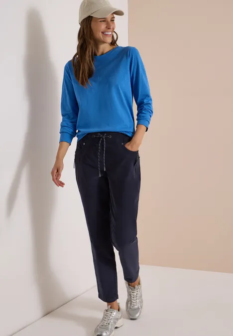 Mid Waist Slim Leg Hose im Casual Fit dark blue corduroy Frau lehnt entspannt an Wand, trägt blaues Sweatshirt, dunkelblaue Hose, weiße Sneaker und weiße Cap.