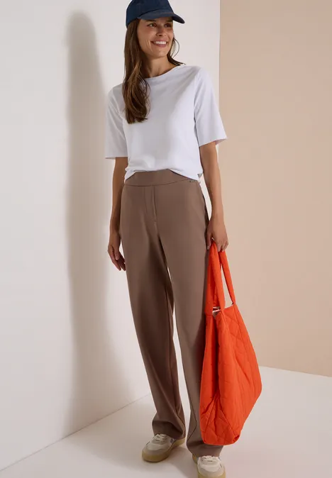 Eine junge Frau in weißem T-Shirt, brauner Hose, blauer Kappe hält eine orange Tasche.