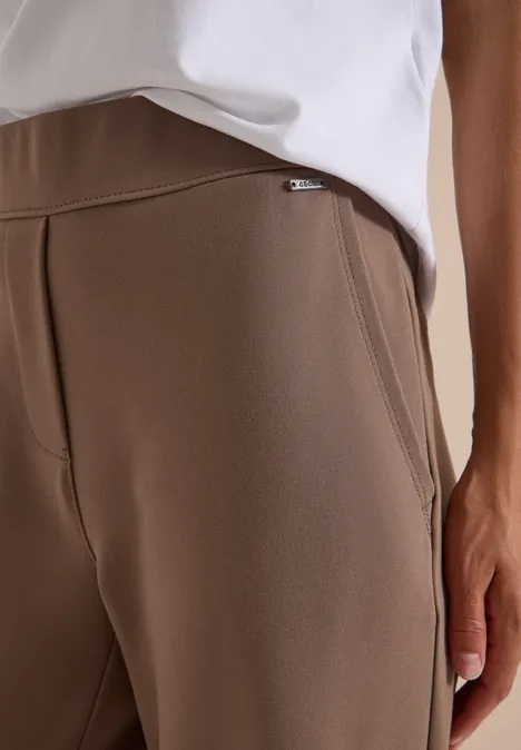 Nahaufnahme einer Person in braunen Stoff-Hose mit Tasche und weißem Oberteil.
