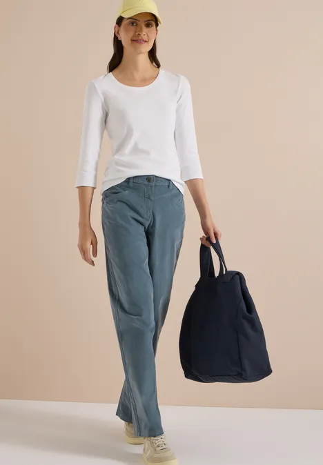 High Waist Wide Leg Hose im Loose Fit dove blue Frau in weißem Langarmshirt, Jeans und Cap trägt eine große dunkelblaue Tasche vor beigem Hintergrund.