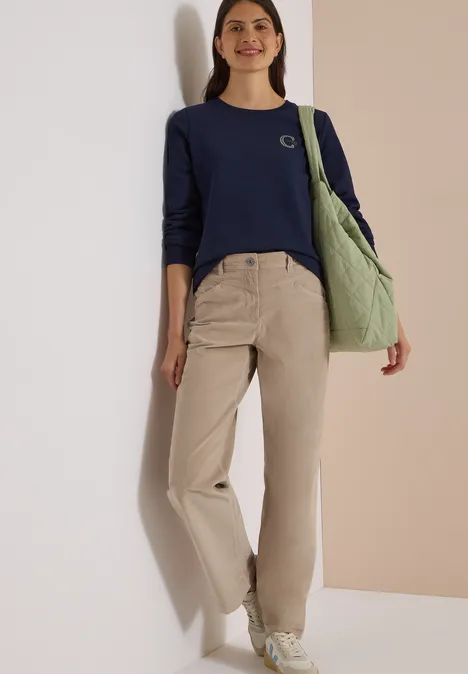 High Waist Wide Leg Hose im Loose Fit silvery beige Frau in navyblauem Pullover, beigen Hosen und weißen Sneakern, trägt grüne Tasche.