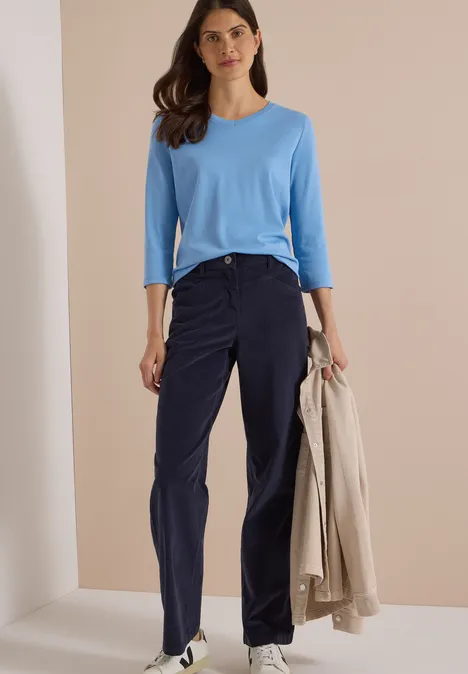 High Waist Wide Leg Hose im Loose Fit dark blue corduroy Frau in hellblauem Oberteil und dunkelblauer Hose hält einen beigen Mantel und steht vor neutralem Hintergrund.
