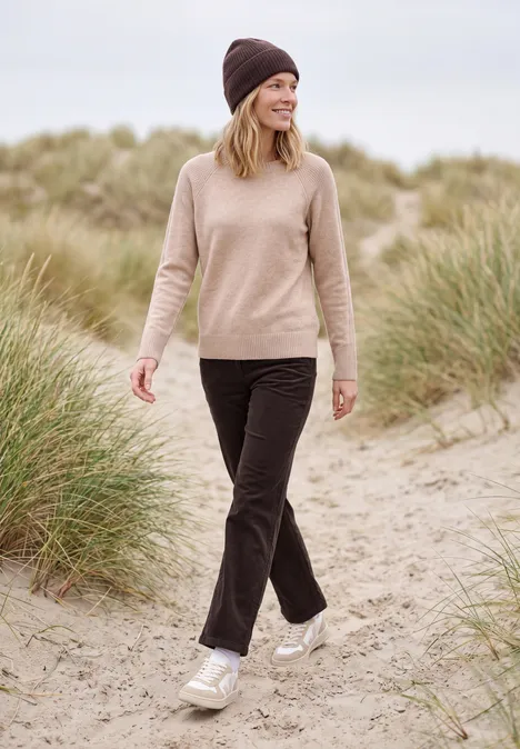 Een vrouw met beige muts en trui loopt op sneakers over zandduinen met helmgras.