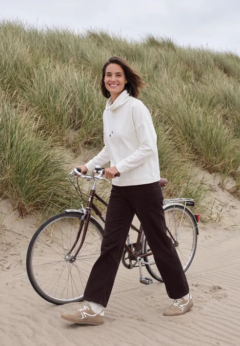 Broek met High Waist en Wide Leg pijpen in een Loose Fit pasvorm berry red Een vrouw in witte trui wandelt lachend met haar fiets langs grasduinen.