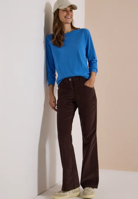 Eine Frau lehnt an einer Wand, trägt ein blaues Shirt, braune Hose und beige Baseballkappe, sie lächelt.