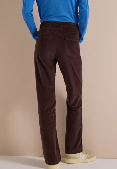 Rückansicht einer Person mit dunkelbraunen Cordhose, blauem Oberteil und hellen Sneakers vor neutralem Hintergrund.