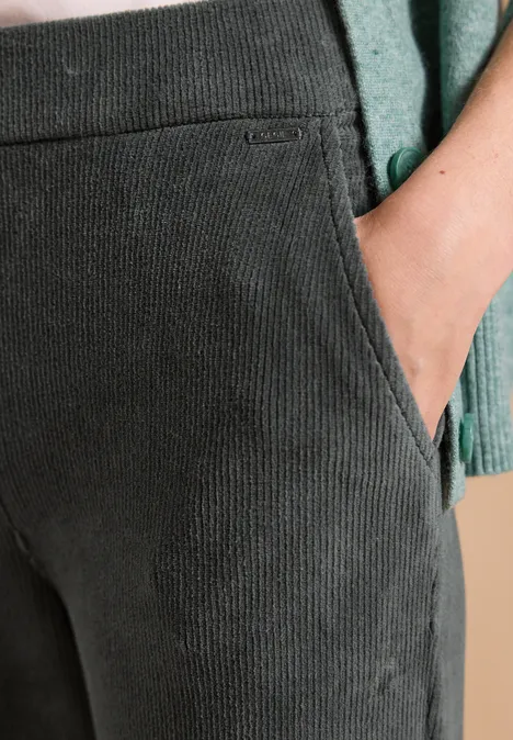 Person trägt graue Cordhose und grünes Oberteil, Hand steckt in der Tasche.