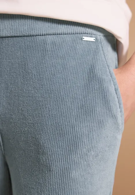 Detail einer grauen, gerippten Hose mit Tasche und kleinem Metalllogo.