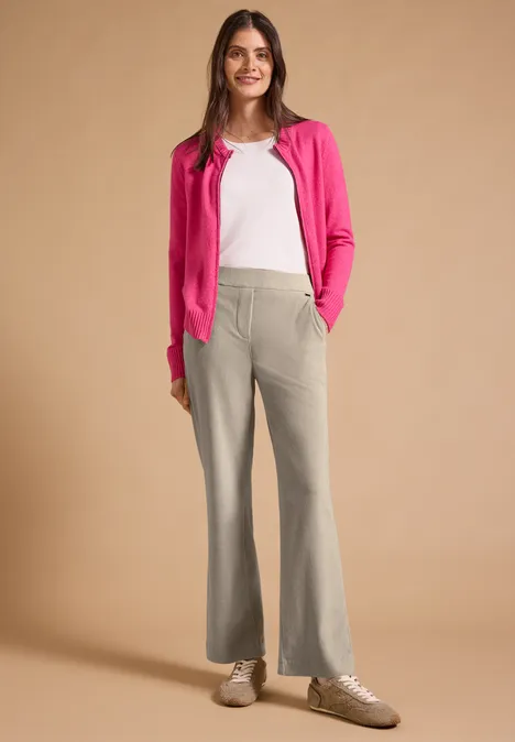 Frau trägt pinken Cardigan, weißes Shirt, graue Schlaghose und Sneakers vor neutralem Hintergrund.