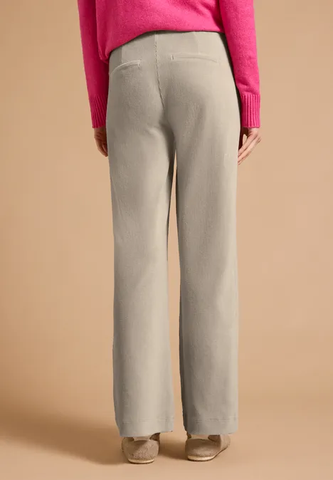 Person von hinten in beige-weiten Hosen, rosa Oberteil und beigen Schuhen vor schlichtem Hintergrund.