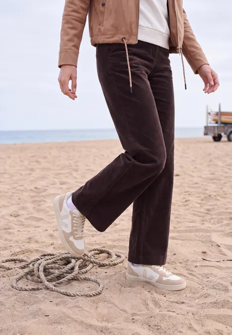 High Waist Straight Leg Hose im Loose Fit berry red Frau in brauner Cord-Hose und weißen Sneakern am Strand stehend neben einem Seil