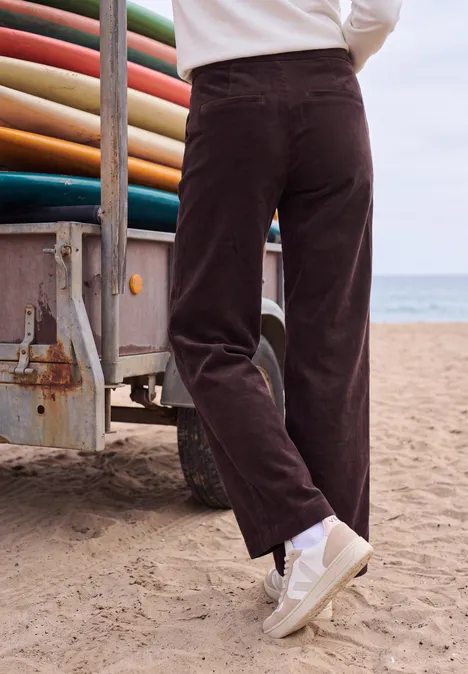 High Waist Straight Leg Hose im Loose Fit berry red Person am Strand trägt braune Cordhose und weiße Sneaker, steht neben Van mit Surfboards.