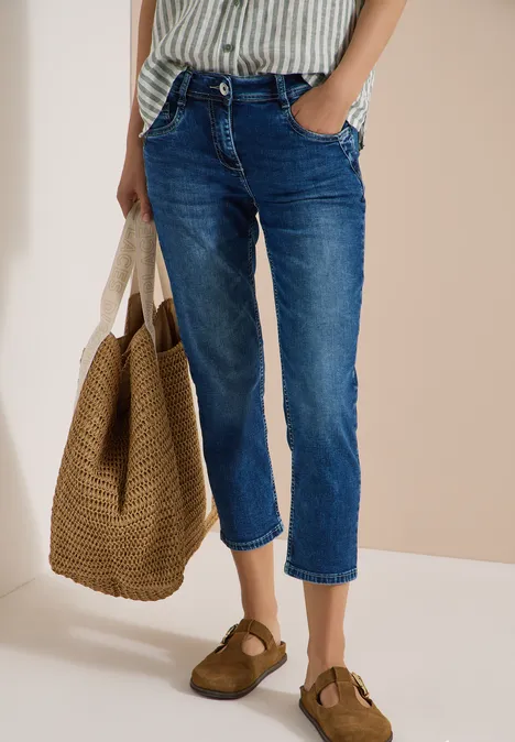 Personne en chemise à fines rayures, jean court bleu, portant un sac en paille et des sandales marron.