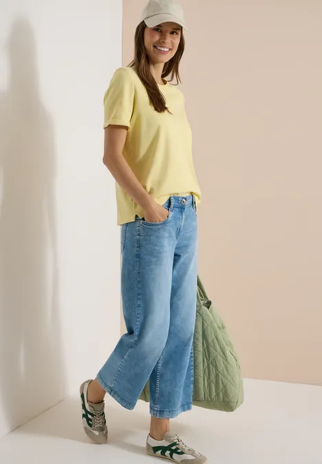 Femme en t-shirt jaune, jean large, sandales et tote bag vert en tissu.