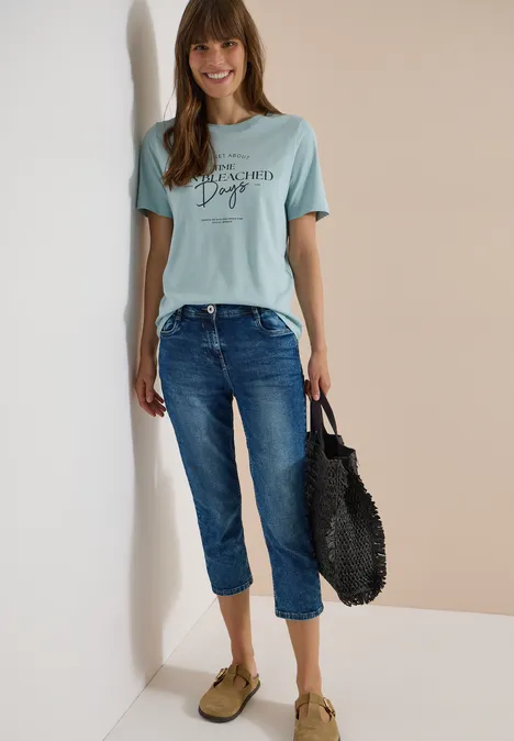 Femme en t-shirt bleu clair, jean décontracté, mules vertes et sac panier noir devant un mur bicolore.