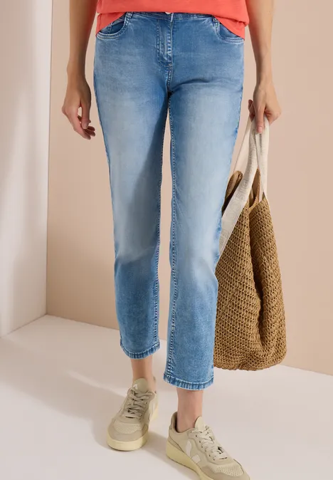 Jean taille mi-haute à coupe Straight Leg et coupe décontractée light blue wash Femme en haut rouge, jean bleu clair, baskets beiges et sac en paille devant un mur clair.