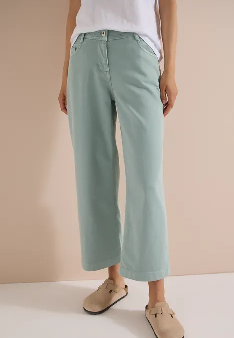 Frau trägt weite, pastellgrüne Hosen und beige Clogs vor neutralem Hintergrund.